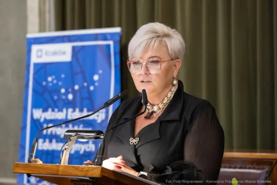 Kobieta w okularach wygłasza przemówienie na tle banera edukacyjnego.