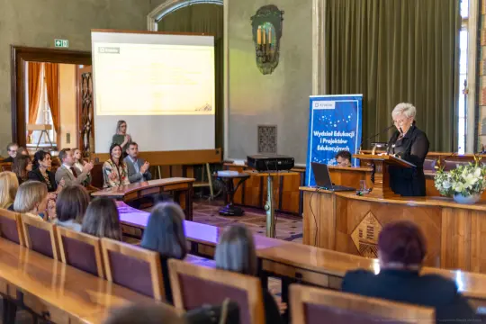 Konferencja edukacyjna z prelegentką przy mównicy i uczestnikami w tle.