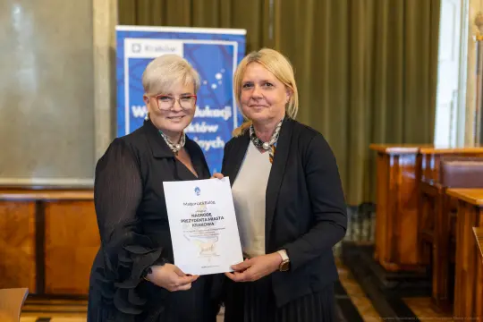 Dwie kobiety trzymają dokument w sali, w tle baner z logo Krakowa.