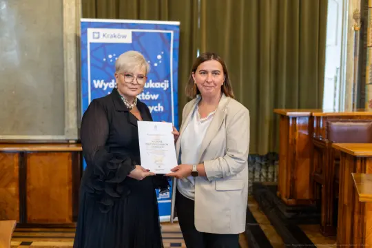 Dwie kobiety wręczają sobie dyplom w sali z drewnianymi elementami.