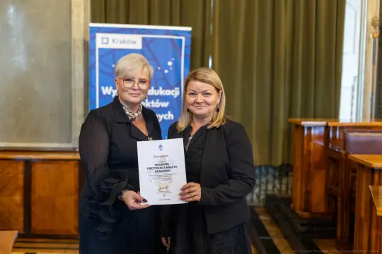 Dwie kobiety trzymają dyplom w sali z drewnianymi elementami.