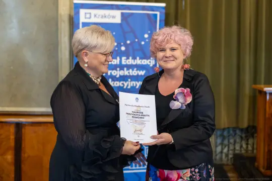 Kobieta wręczająca dyplom drugiej kobiecie w eleganckim otoczeniu.