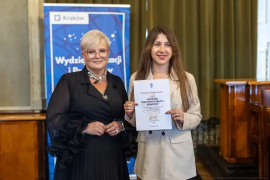 Kobieta z dyplomem i druga w eleganckim stroju, obie w sali z dekoracjami.