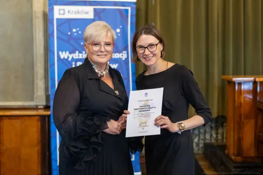 Dwie kobiety trzymają dyplom, uśmiechając się do kamery w eleganckim wnętrzu.