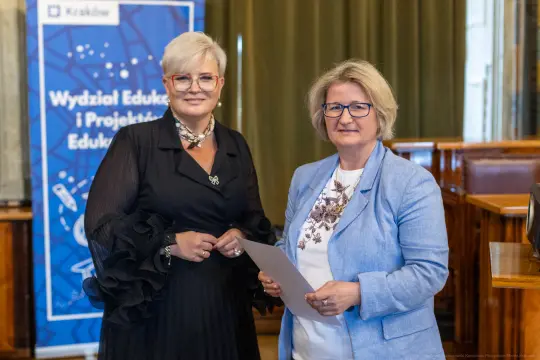 Dwie kobiety w formalnych strojach, uśmiechające się i trzymające dokumenty.