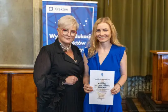 Dwie kobiety trzymają dyplom w sali z zielonymi zasłonami.