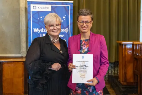 Dwie kobiety trzymają dyplom w sali z eleganckim wystrojem.
