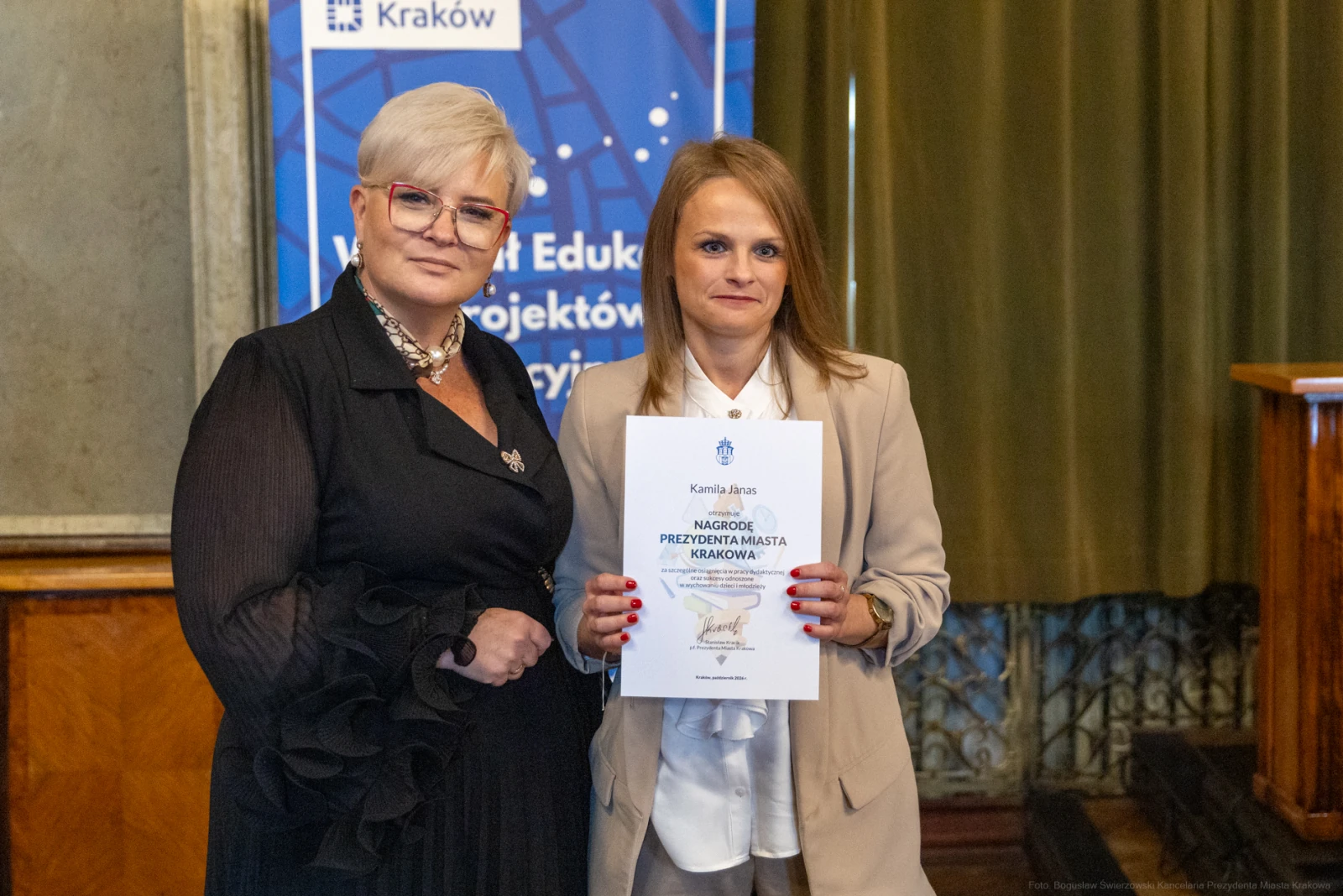 Kobieta wręczająca dyplom drugiej kobiecie w eleganckim otoczeniu.  Autor: Bogusław Świerzowski
