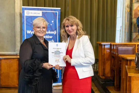 Dwie kobiety trzymają dyplom w sali z dekoracjami.