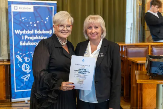 Dwie kobiety trzymają dyplom w sali z dekoracjami edukacyjnymi.