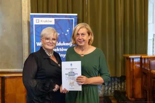 Dwie kobiety trzymają dyplom w sali z zielonymi zasłonami.