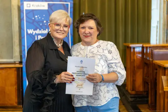 Kobieta wręczająca dyplom drugiej kobiecie w formalnym otoczeniu.