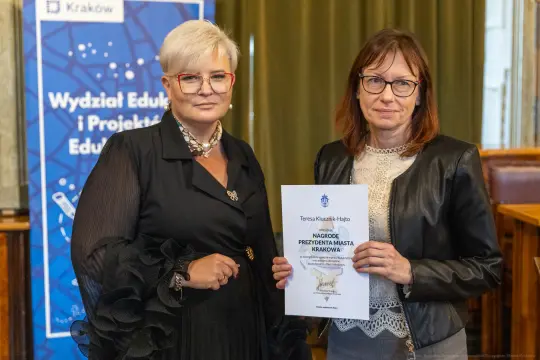 Dwie kobiety trzymają dyplom w sali z dekoracjami, jedna z nich w okularach.