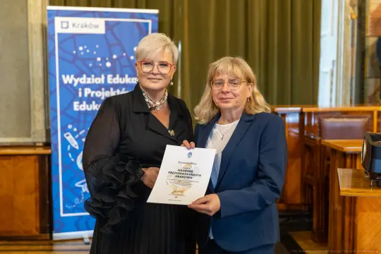 Dwie kobiety trzymają dyplom w sali z banerem Wydziału Edukacji i Projektów Edukacyjnych.