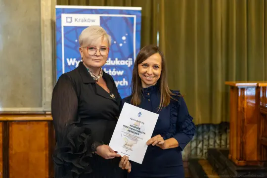 Dwie kobiety trzymają dyplom w sali, jedna z nich w okularach, druga w granatowej bluzce.