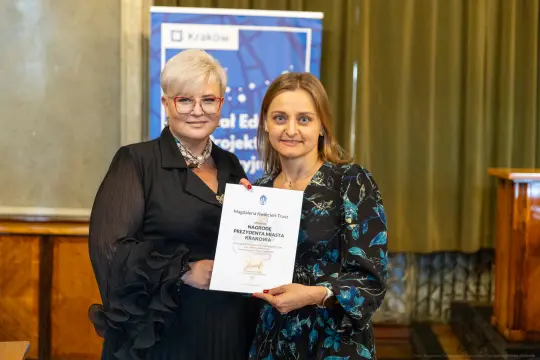 Dwie kobiety trzymają dyplom w sali z dekoracjami.