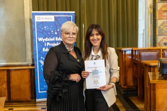 Dwie kobiety trzymają dyplom w sali z drewnianymi elementami i plakatem w tle.