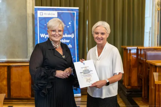 Dwie kobiety trzymają dyplom w sali z drewnianymi elementami.