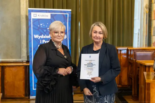 Dwie kobiety trzymają dokument w sali z zielonymi zasłonami.