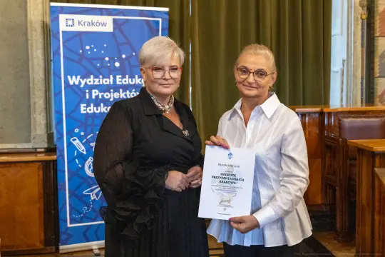 Dwie kobiety trzymają dyplom w sali z dekoracjami i plakatem Wydziału Edukacji.