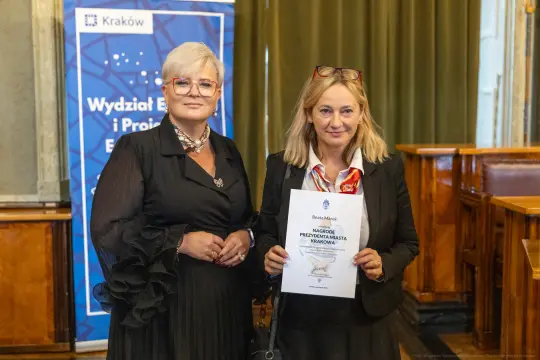 Dwie kobiety w eleganckich strojach trzymają dyplom w sali konferencyjnej.
