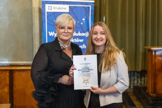 Dwie kobiety trzymają dyplom w sali z zielonymi zasłonami.