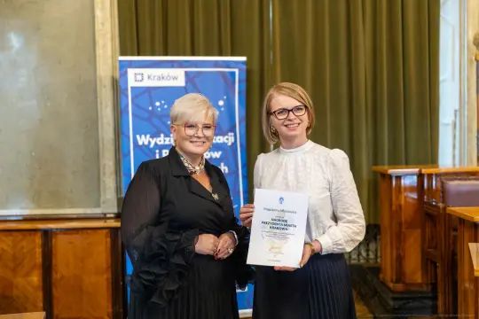 Dwie kobiety trzymają dyplom w sali z zielonymi zasłonami.