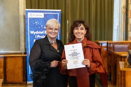Dwie kobiety trzymają certyfikat w sali z drewnianymi elementami.