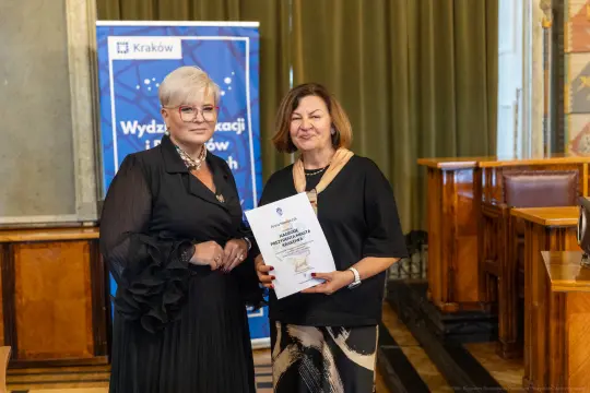 Dwie kobiety trzymające dyplom w sali z dekoracjami.