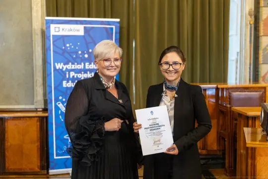Dwie kobiety trzymają dokument w sali konferencyjnej. W tle baner Wydziału Edukacji i Projektów Edukacyjnych.