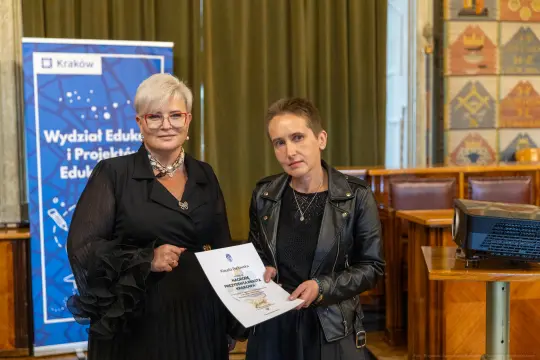 Dwie kobiety trzymają dokument w sali z dekoracyjnymi ścianami.