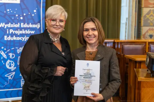 Dwie kobiety trzymają dyplom w sali z dekoracjami.