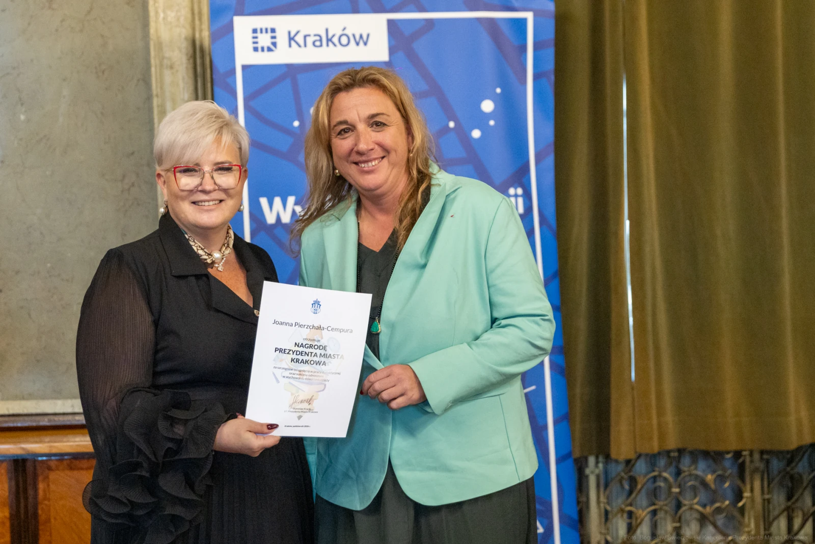 Dwie kobiety trzymają dyplom w sali z dekoracjami, w tle logo Krakowa.  Autor: Bogusław Świerzowski