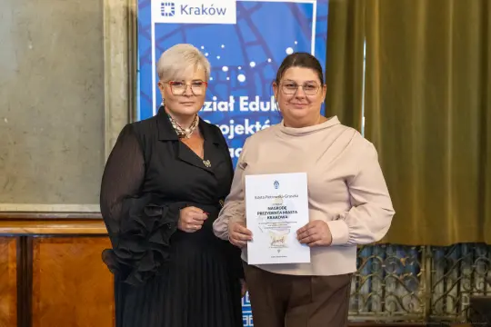 Dwie kobiety trzymające dyplom w sali z dekoracjami.