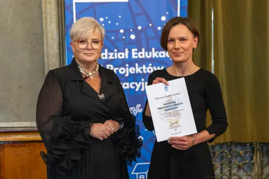 Dwie kobiety trzymają certyfikat w sali z dekoracjami.
