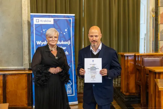 Wręczenie dyplomu w sali z osobą w eleganckim stroju i mężczyzną trzymającym certyfikat.