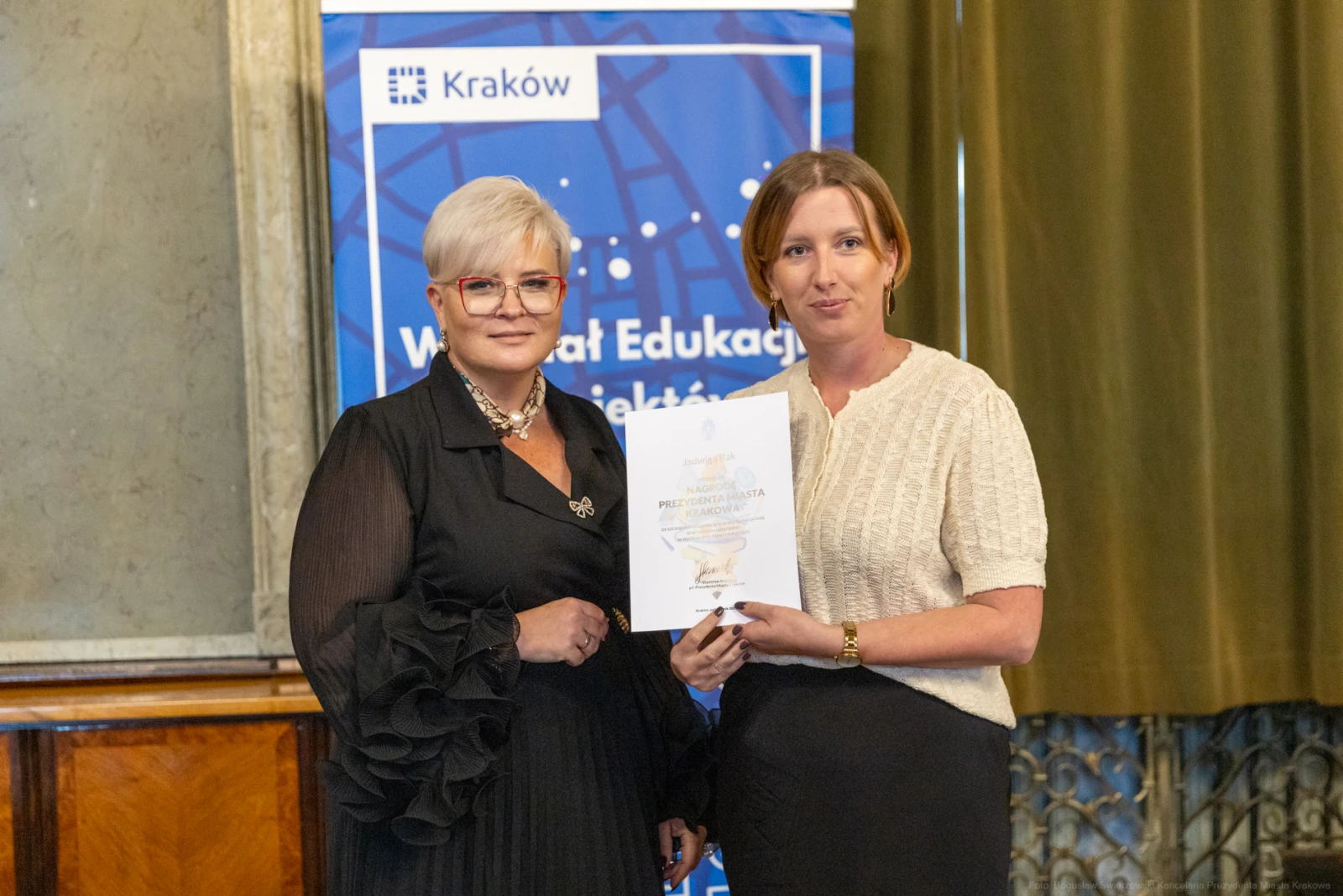 Dwie kobiety trzymają dyplom w sali z dekoracjami. W tle baner z napisem Kraków.  Autor: Bogusław Świerzowski
