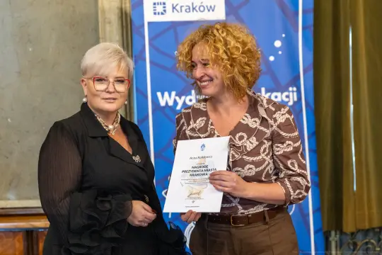 Dwie kobiety uśmiechają się, jedna wręcza drugiej dyplom nagrody.