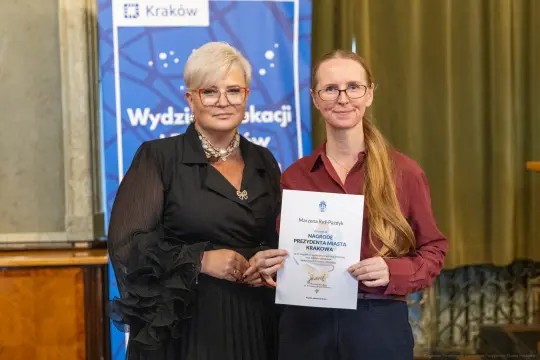 Dwie kobiety trzymają dyplom w sali z zielonymi zasłonami.