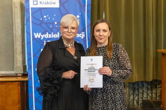 Kobieta wręczająca dyplom drugiej kobiecie w eleganckim otoczeniu.