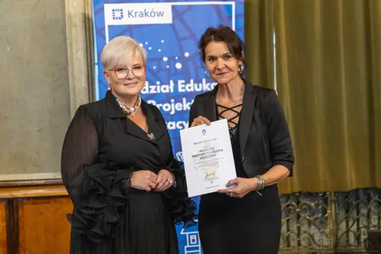 Dwie kobiety trzymające dyplom w sali z dekoracjami.