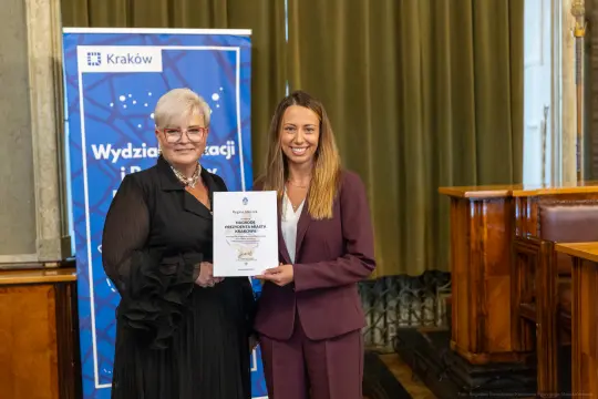 Dwie kobiety trzymają dyplom w sali z zielonymi zasłonami.