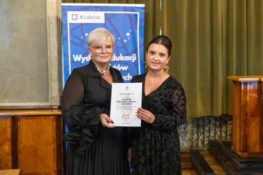 Dwie kobiety trzymają dokument w sali z drewnianymi elementami i banerem w tle.