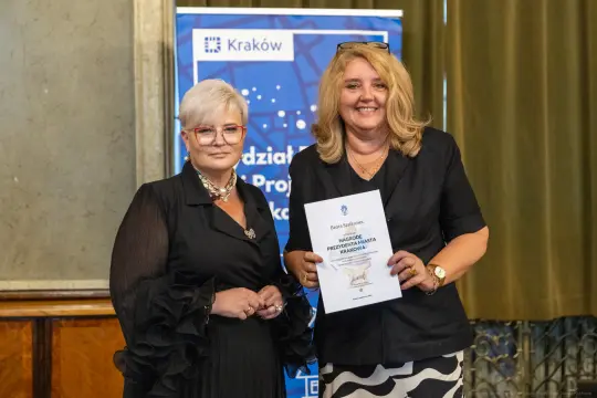 Dwie kobiety trzymają dokument, uśmiechając się w formalnym otoczeniu.