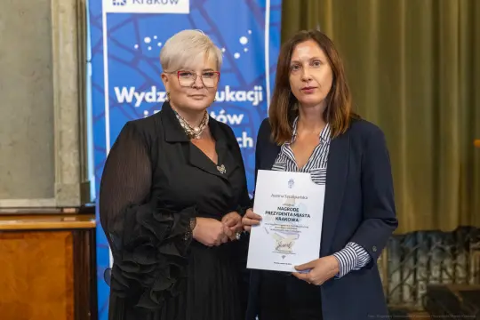 Dwie kobiety trzymające dyplom w sali z zielonymi zasłonami.