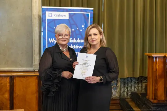 Dwie kobiety trzymające dyplom w sali z zielonymi zasłonami.