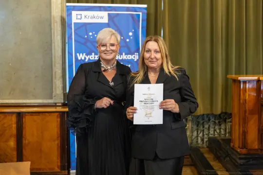 Dwie kobiety trzymają dyplom w sali z zielonymi zasłonami i banerem Wydziału Edukacji.