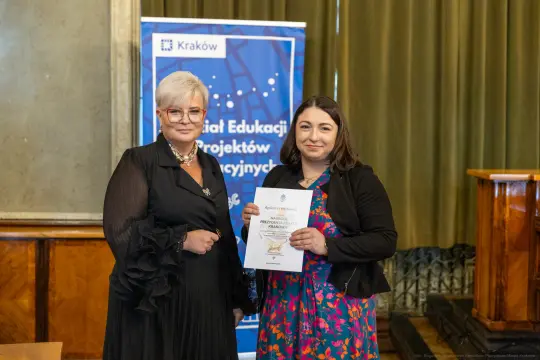 Dwie kobiety trzymają dyplom w sali z zielonymi zasłonami.