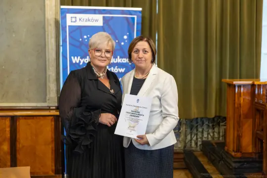 Dwie kobiety trzymające dokument w sali z zielonymi zasłonami.