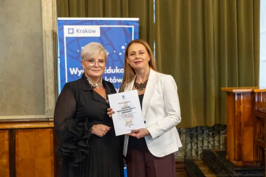 Dwie kobiety wręczają sobie dyplom w sali z zielonymi zasłonami.